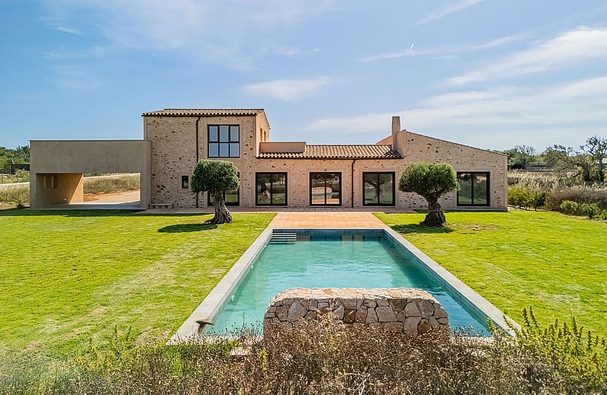 Exterior view of a stone finca in Santanyí featuring a rectangular swimming pool, manicured lawn, and large glass doors.