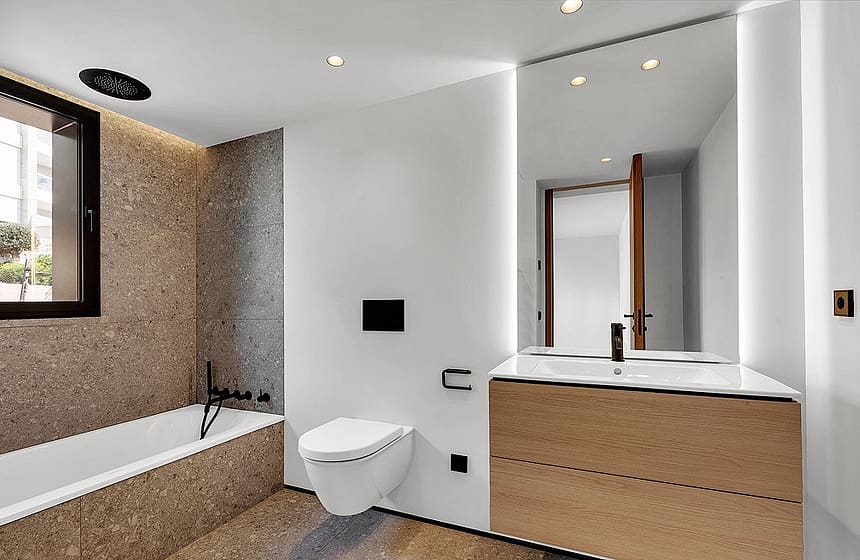 Bathroom interior featuring a wall-mounted toilet, light wood vanity, and brown stone tile bathtub surround with matte black fixtures.