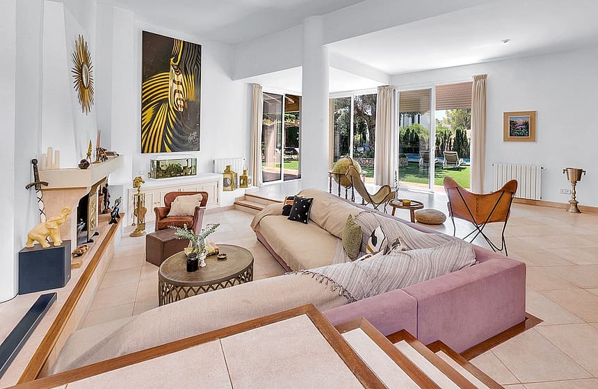 A bright, open-plan living room with a large corner sofa, a fireplace, and glass doors leading to an outdoor garden area.