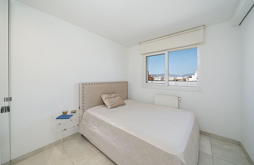 A minimalist bedroom featuring a neutral upholstered bed, white walls, and a window framing an urban Old Town vista.