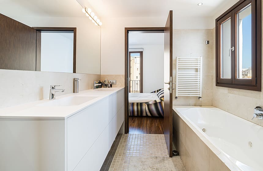 Bright bathroom featuring a white double vanity, integrated bathtub, natural stone wall tiling, and wooden door access.