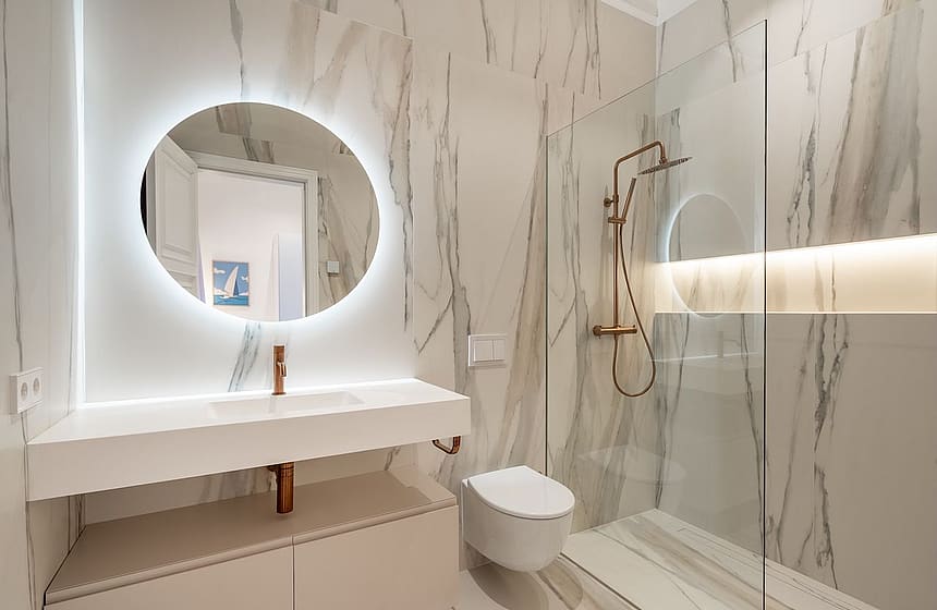 A modern bathroom featuring white marble wall tiles, a floating vanity, a backlit circular mirror, and a glass-enclosed shower.