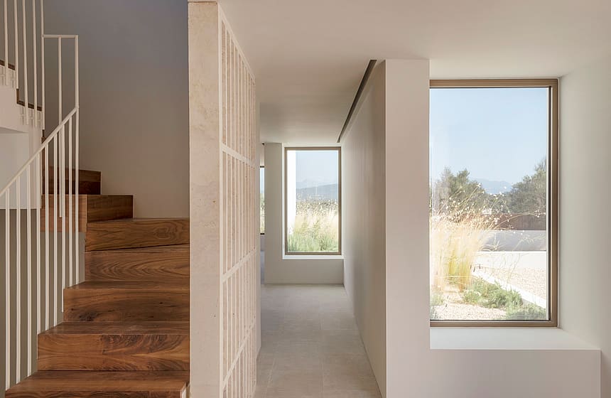 A bright minimalist hallway featuring a vertical timber screen, wooden stairs, and large windows overlooking a field.