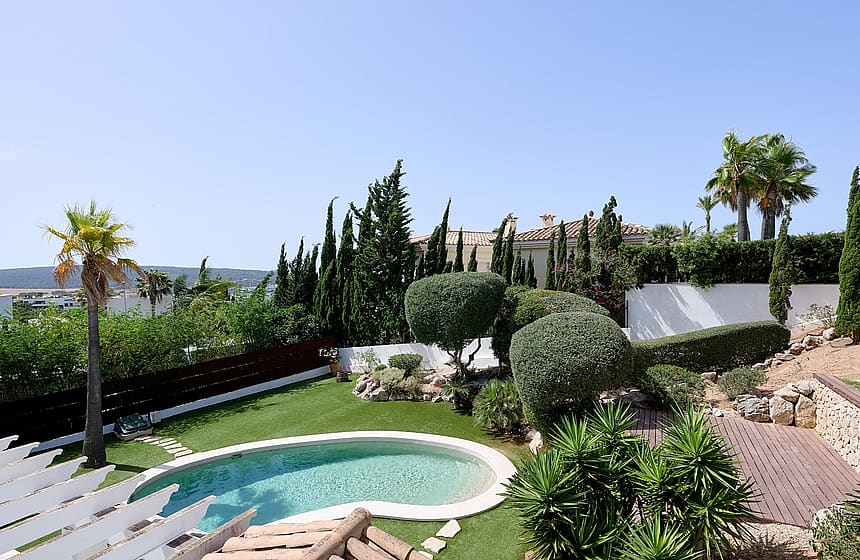Vista desde tarima de madera sobre piscina ovalada, césped artificial, topiaria y palmeras con montañas al fondo