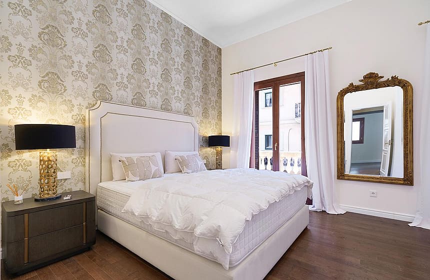 A master bedroom with a large bed against a damask patterned wall, dark wood floors, and a floor-length gold-framed mirror.