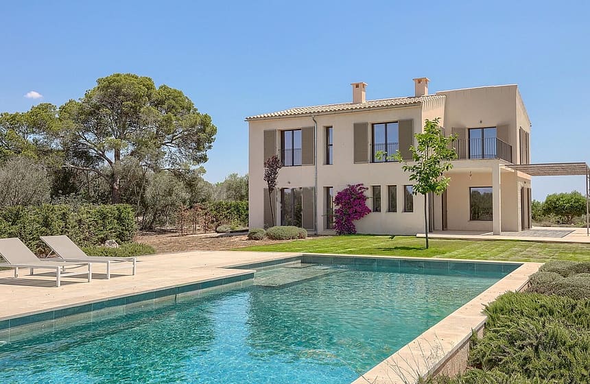 Exterior view of a beige stone finca with wooden shutters, a swimming pool, and lounge chairs in a landscaped garden.