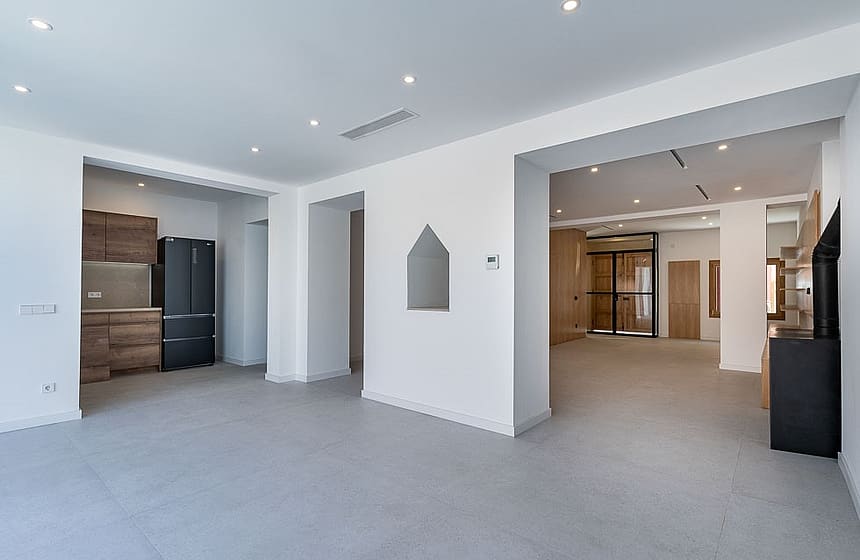 Wide shot of a contemporary interior with an open kitchen featuring dark wood cabinetry and a large refrigerator, transitioning to a bright, minimalist living space with grey tiled flooring and recessed ceiling lights.