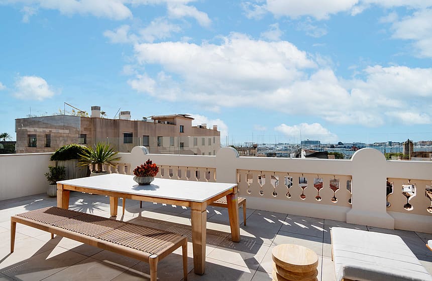 Rooftop terrace featuring a light wooden dining table with a bench, beige stone tiling, and traditional balustrade railings overlooking the Santa Catalina urban skyline.