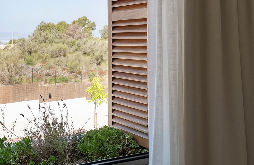 White curtains and wooden louvered shutters frame a window with an integrated planter box and green landscape beyond.