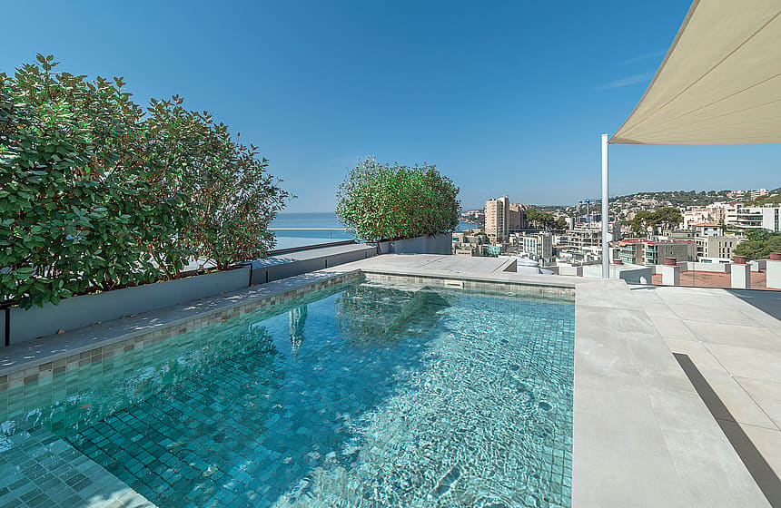A private rooftop plunge pool with light blue tiled interior, framed by green shrubs, overlooking the Mediterranean sea.