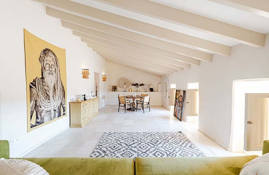 A spacious living room in a Sencelles townhouse featuring high vaulted ceilings with exposed cream beams, white walls, and a large artistic wall hanging.