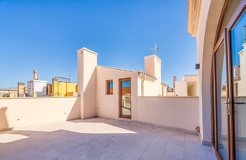An open stone-paved rooftop terrace with light-colored walls and glass doors under a clear blue sky, overlooking city buildings.
