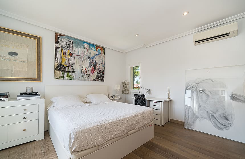 A bright bedroom with white walls, a king-size bed, a large abstract painting, and light-toned hardwood flooring.