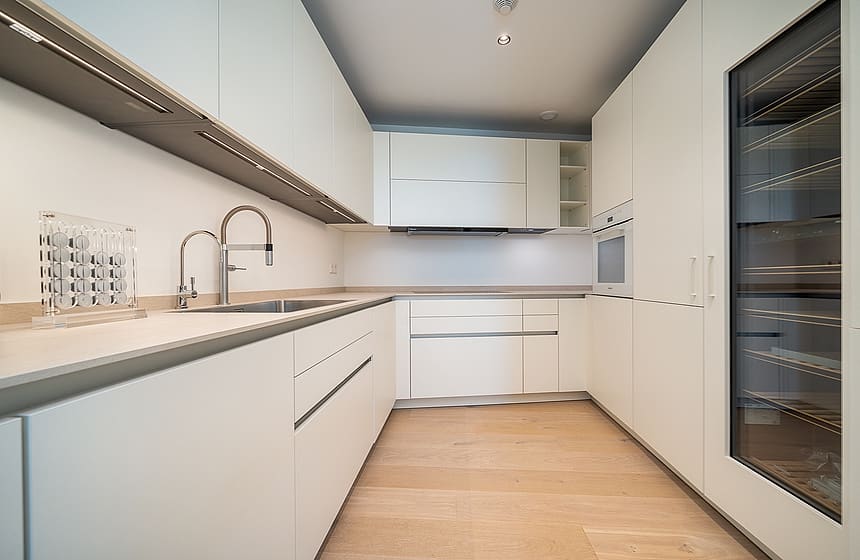A sleek galley-style kitchen with white handleless cabinetry, light oak wood flooring, and a glass-fronted wine refrigerator.
