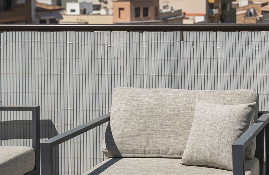 A modern outdoor lounge chair with grey fabric cushions sits on a terrace with a metal privacy screen overlooking a city.