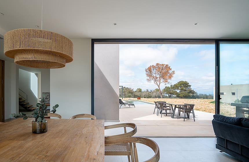 Dining area with a large wooden table and textured pendant light opening onto a terrace with a stone wall and mountain views.