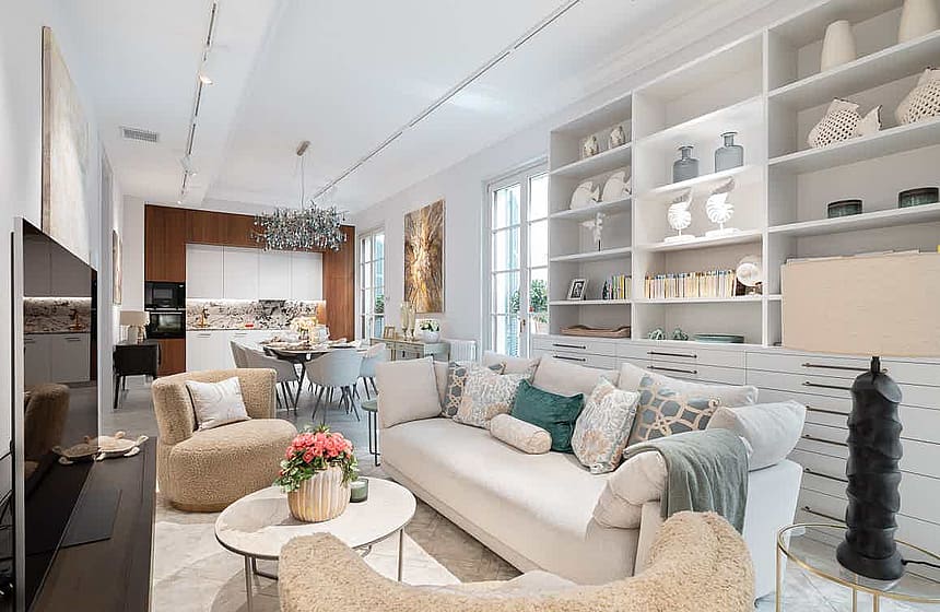 Open-plan living room with white custom built-in shelving, a neutral beige sofa, and two plush armchairs facing a kitchen area.