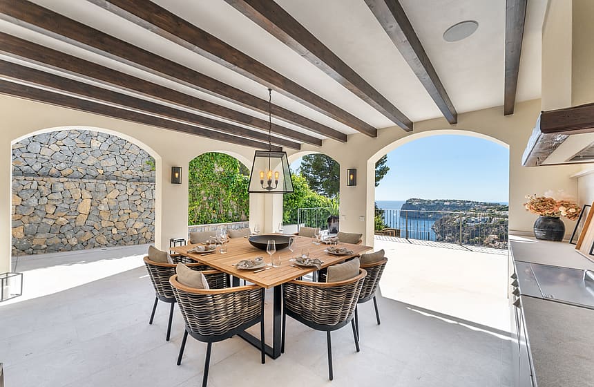 Covered terrace with dark wooden ceiling beams, a large square table, woven chairs, and views of a sea bay through arches.