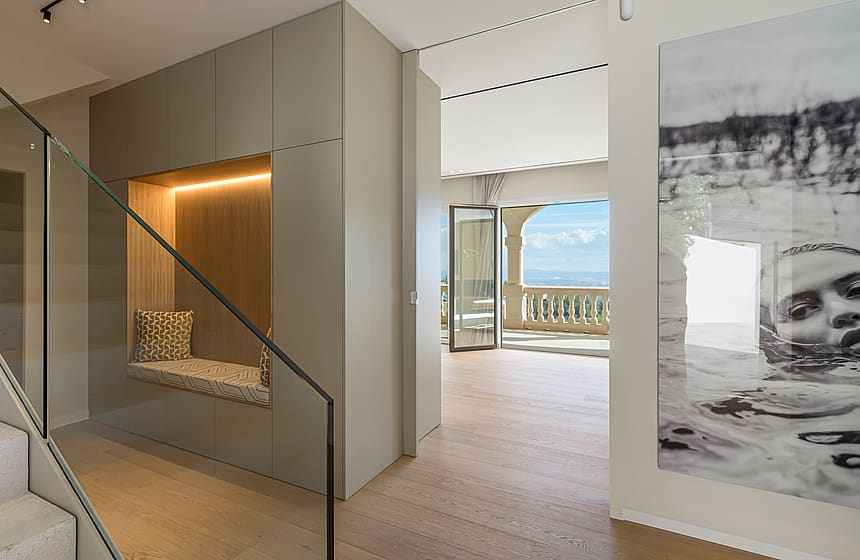 A modern staircase with a frameless glass railing leads past a built-in wall niche with a padded bench and warm lighting.