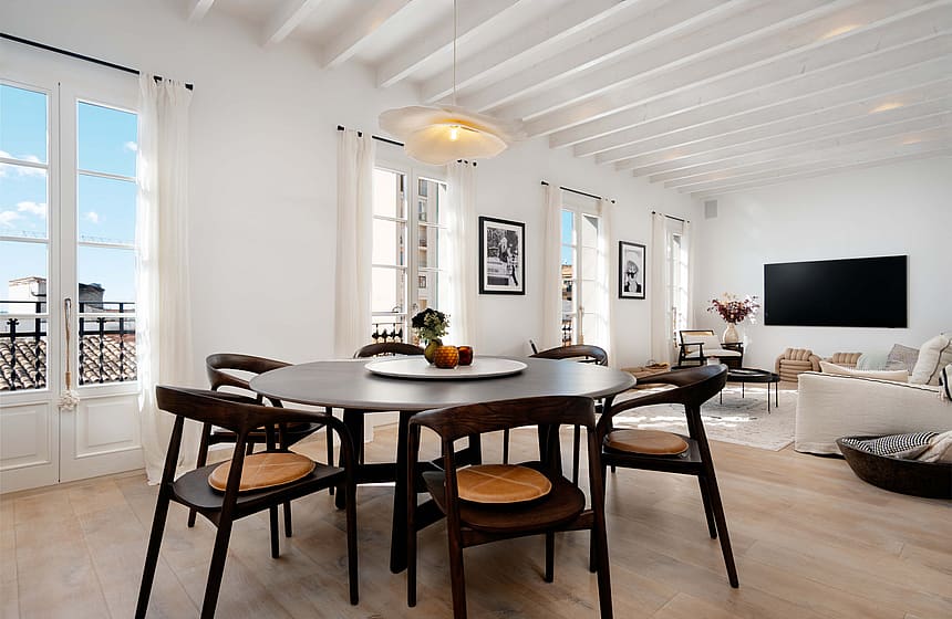 An airy, sunlit open-plan room with a round dark wood dining table, white beamed ceilings, and floor-to-ceiling French doors.