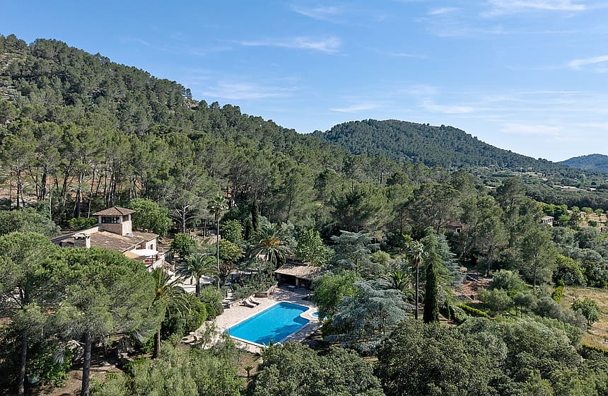 Aerial perspective of a stone finca surrounded by pine forests, showing a swimming pool and terraced gardens in Alaro.