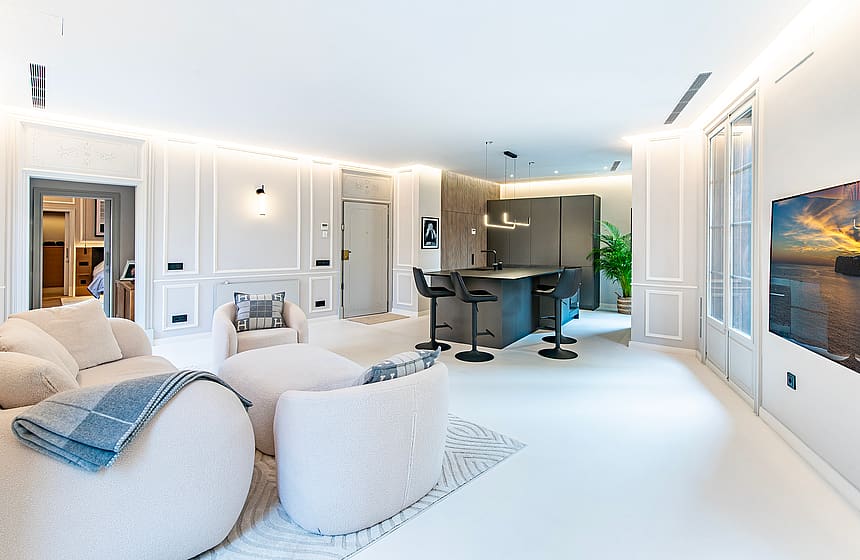 Wide shot of a modern white open-plan living room with a kitchen island, textured armchairs, and floor-to-ceiling windows.