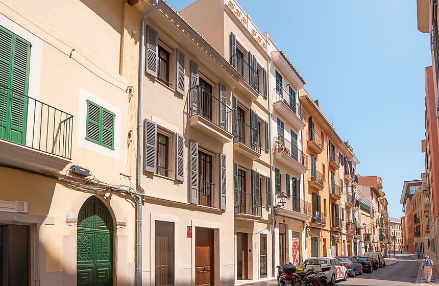 Multi-story Mediterranean townhouses featuring sand-colored plaster, green wooden shutters, and wrought-iron balconies.
