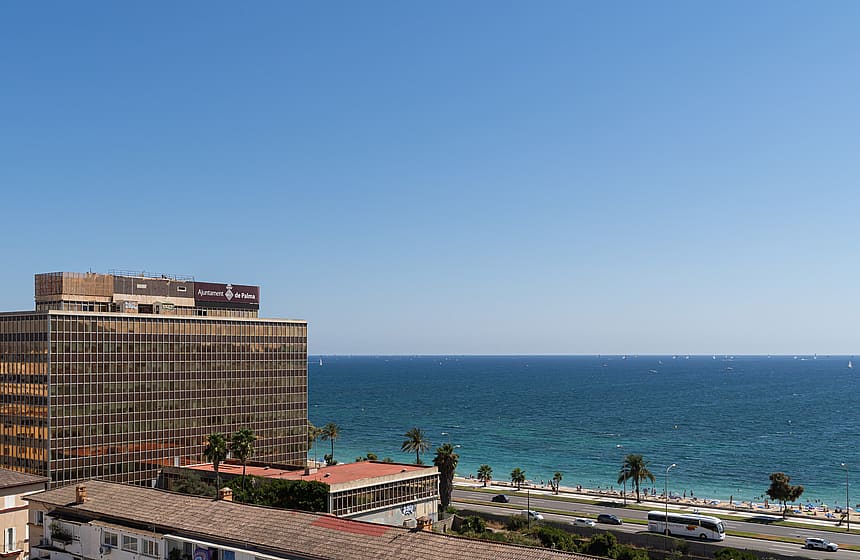 High-angle view overlooking a modern commercial building with mirrored glass facade, a sandy beach, and the Mediterranean Sea.