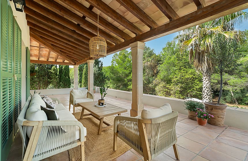 A covered terrace with a timber-beamed ceiling, stone pillars, and neutral lounge furniture overlooking a forest.