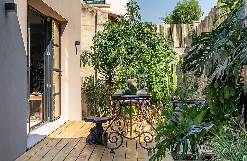 A small outdoor garden space with light-colored wood decking, a wrought iron table, and various potted green plants.