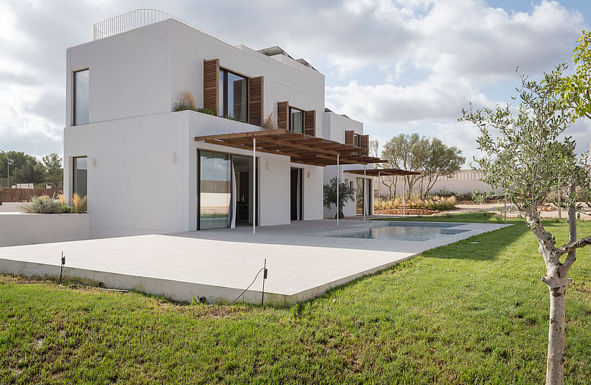 Modern two-story white villa with wooden shutters, a large pergola, and stone terrace overlooking a green lawn.