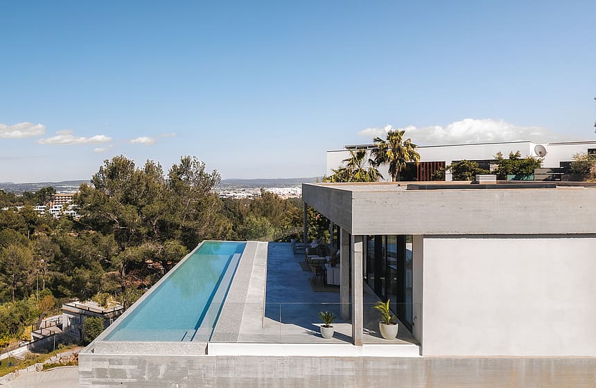 Elevated view of a long infinity lap pool on a grey stone terrace of a minimalist concrete villa overlooking the valley.