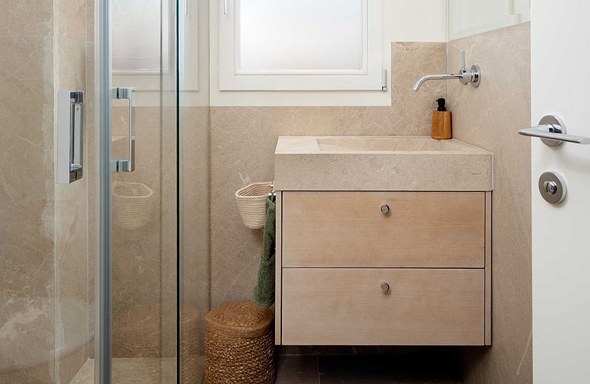A modern small bathroom featuring a floating wooden vanity, a glass-enclosed walk-in shower, and beige stone wall tiling.