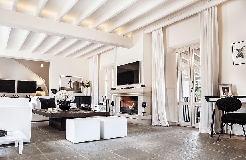 Bright living area with a stone fireplace, exposed beam ceiling, stone flooring, and white linen curtains.
