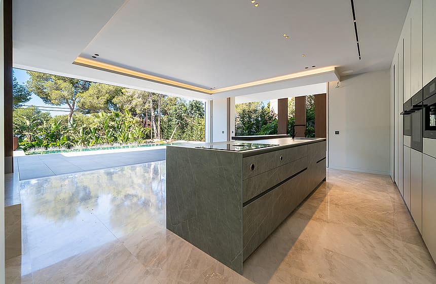 Modern kitchen with a large dark stone island, pale marble floors, and wide sliding doors opening to a garden terrace.