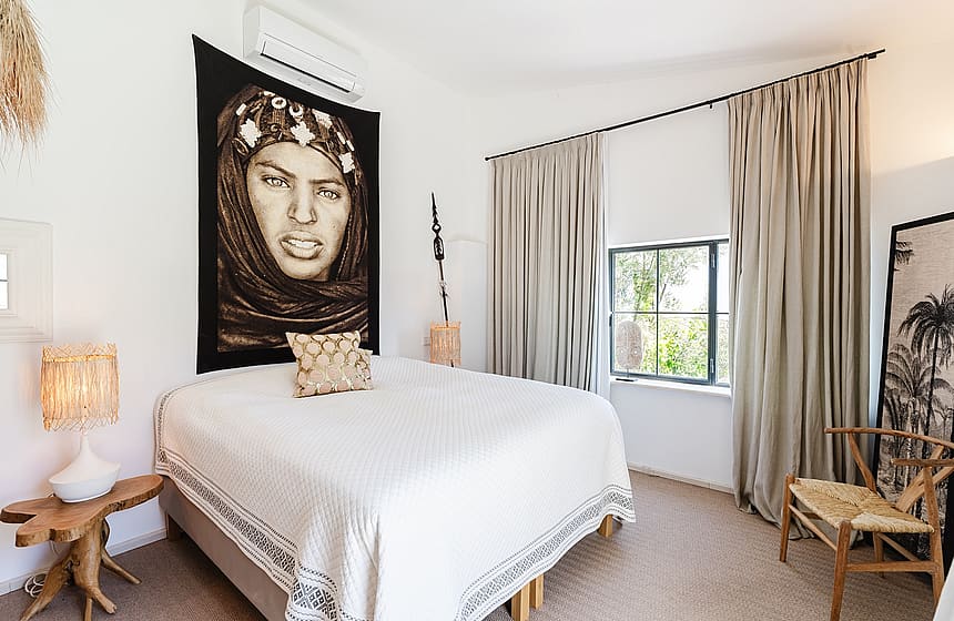 A bedroom featuring a bed with a white quilt, a large decorative wall tapestry above the headboard, beige floor-to-ceiling curtains, and a natural wood side table.
