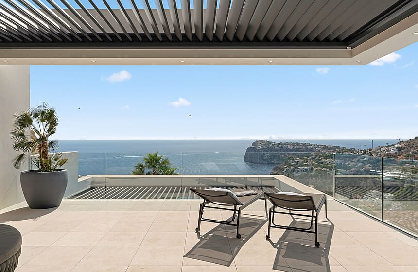 A covered terrace with two sun loungers facing the sea in Port d'Andratx, featuring a glass railing and a louvered pergola.