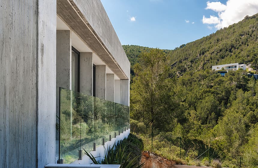 Side profile of a textured concrete villa exterior with glass railings overlooking green hills under a blue sky.