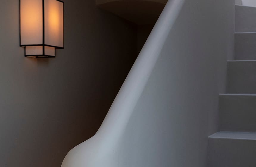 A curved plaster staircase with a modern black lantern wall sconce emitting a warm glow in a minimalist hallway.