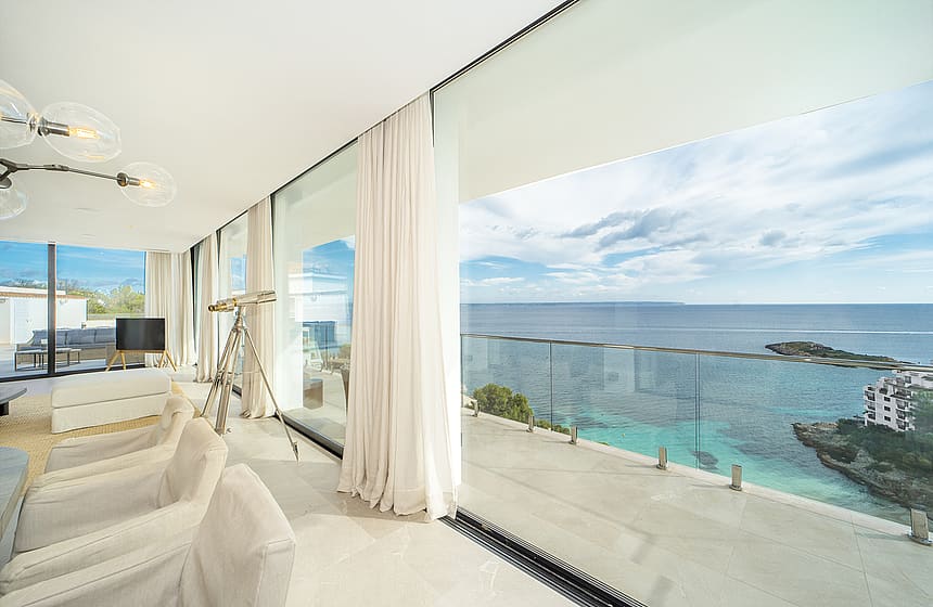 Modern living room with floor-to-ceiling sliding glass doors opening to a terrace, light beige furniture, and sea views.