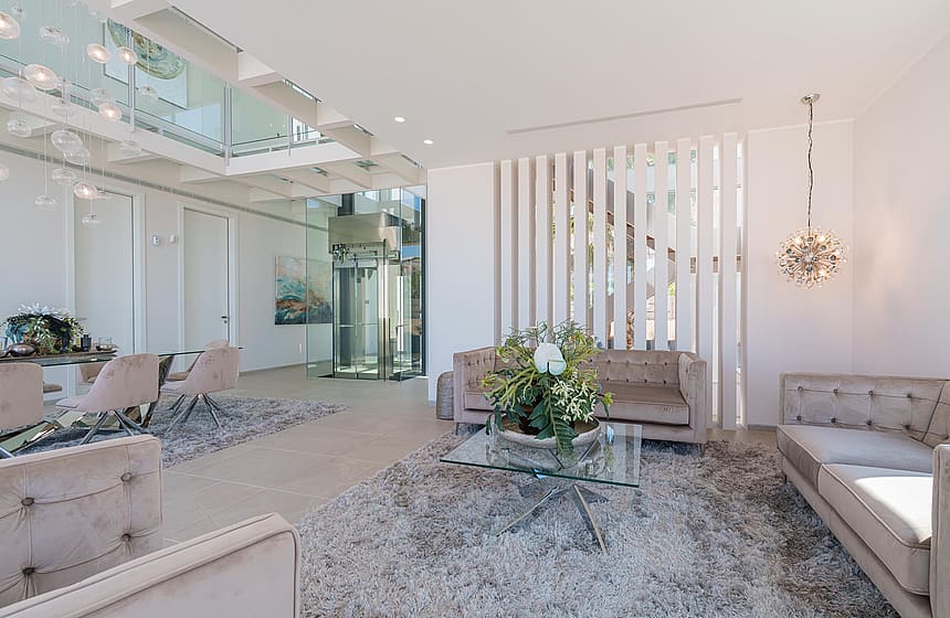 Contemporary living room featuring a central glass elevator, light beige sofas, a textured area rug, and vertical slat partition.