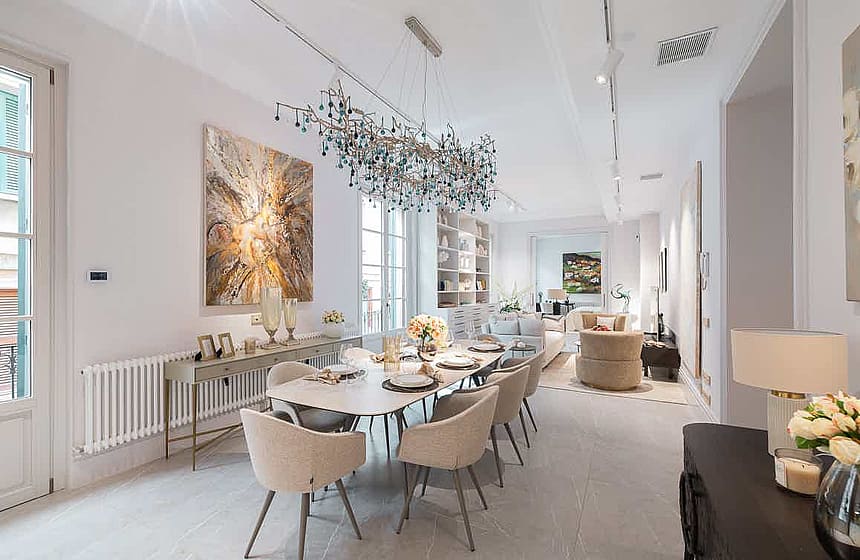 Dining area with a rectangular light-wood table, cream upholstered chairs, and a large sculptural chandelier overhead.