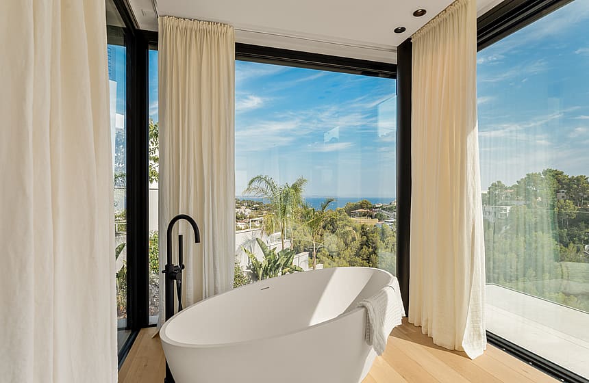 Freestanding white bathtub in a bathroom with floor-to-ceiling windows, cream curtains, and light wood flooring.