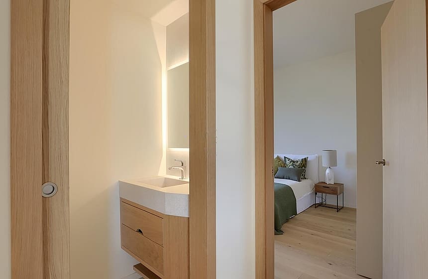 A view through a wooden door frame into a bedroom with a view of a minimalist wooden vanity unit in the foreground.