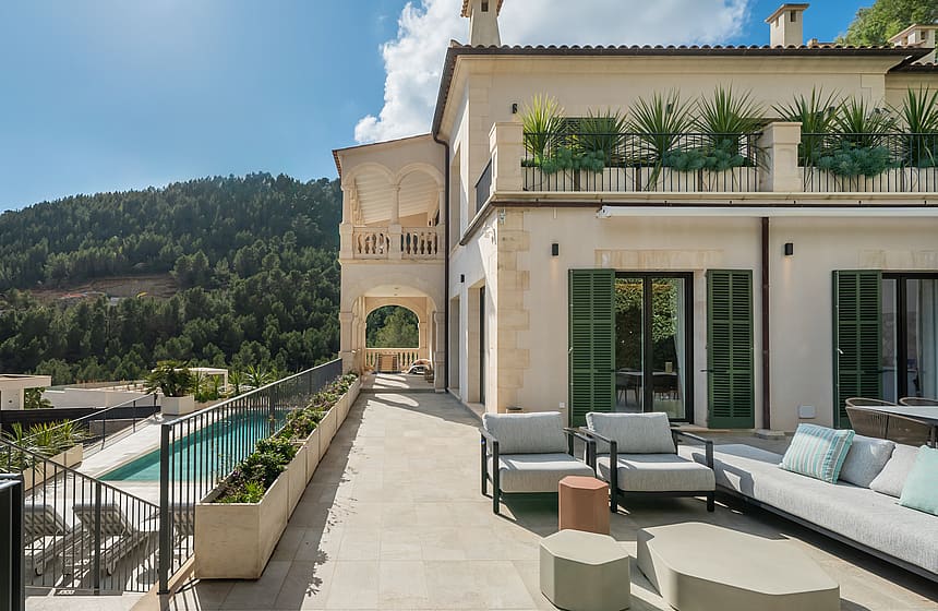 Exterior facade of a cream stone villa with a long swimming pool and shaded outdoor seating area overlooking hills.