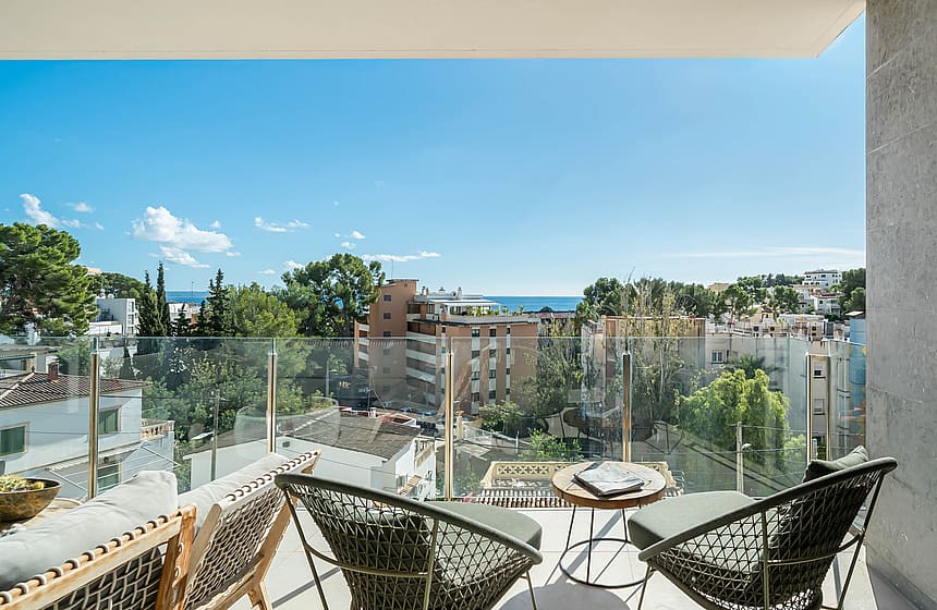 A balcony view featuring two dark wicker armchairs and a small round wooden table overlooking a coastal town and the sea.