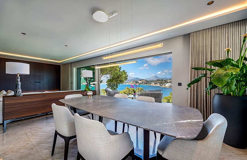Dining room with a large oval grey stone table and upholstered chairs, facing an open terrace overlooking a blue bay.