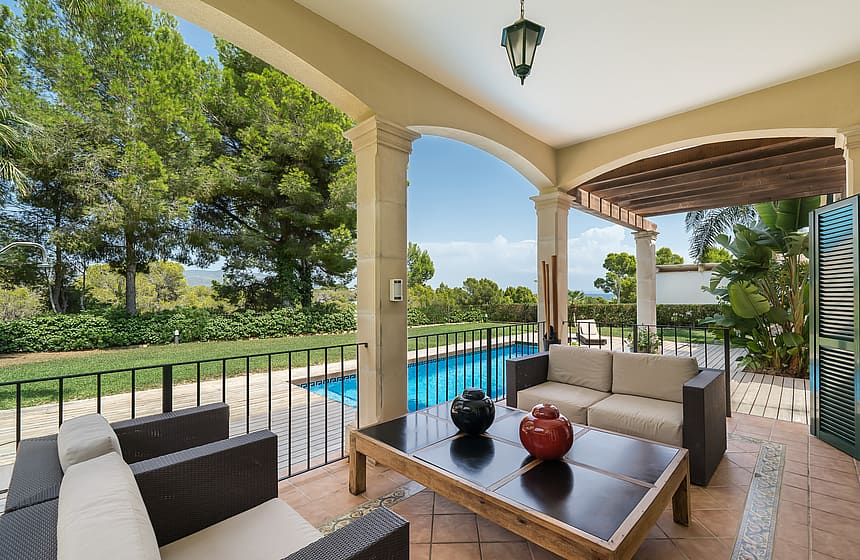 A covered terrace with comfortable outdoor seating overlooks a blue swimming pool and surrounding green gardens on a sunny day.