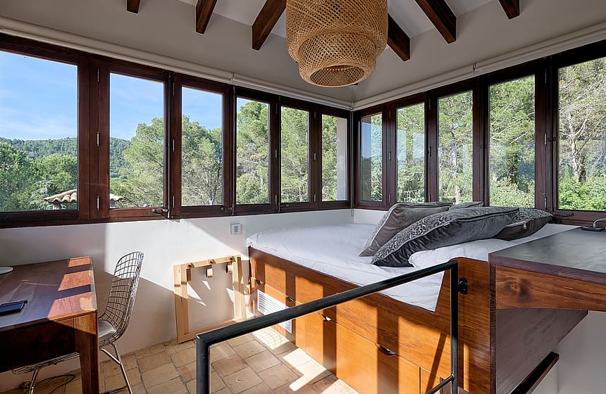 A sunlit bedroom in a finca with wooden ceiling beams, large windows overlooking forest, and a built-in wooden bed frame.