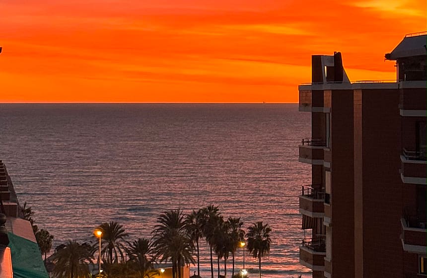 A vibrant orange sunset over the sea viewed from an elevated position, with silhouettes of buildings and palm trees below.